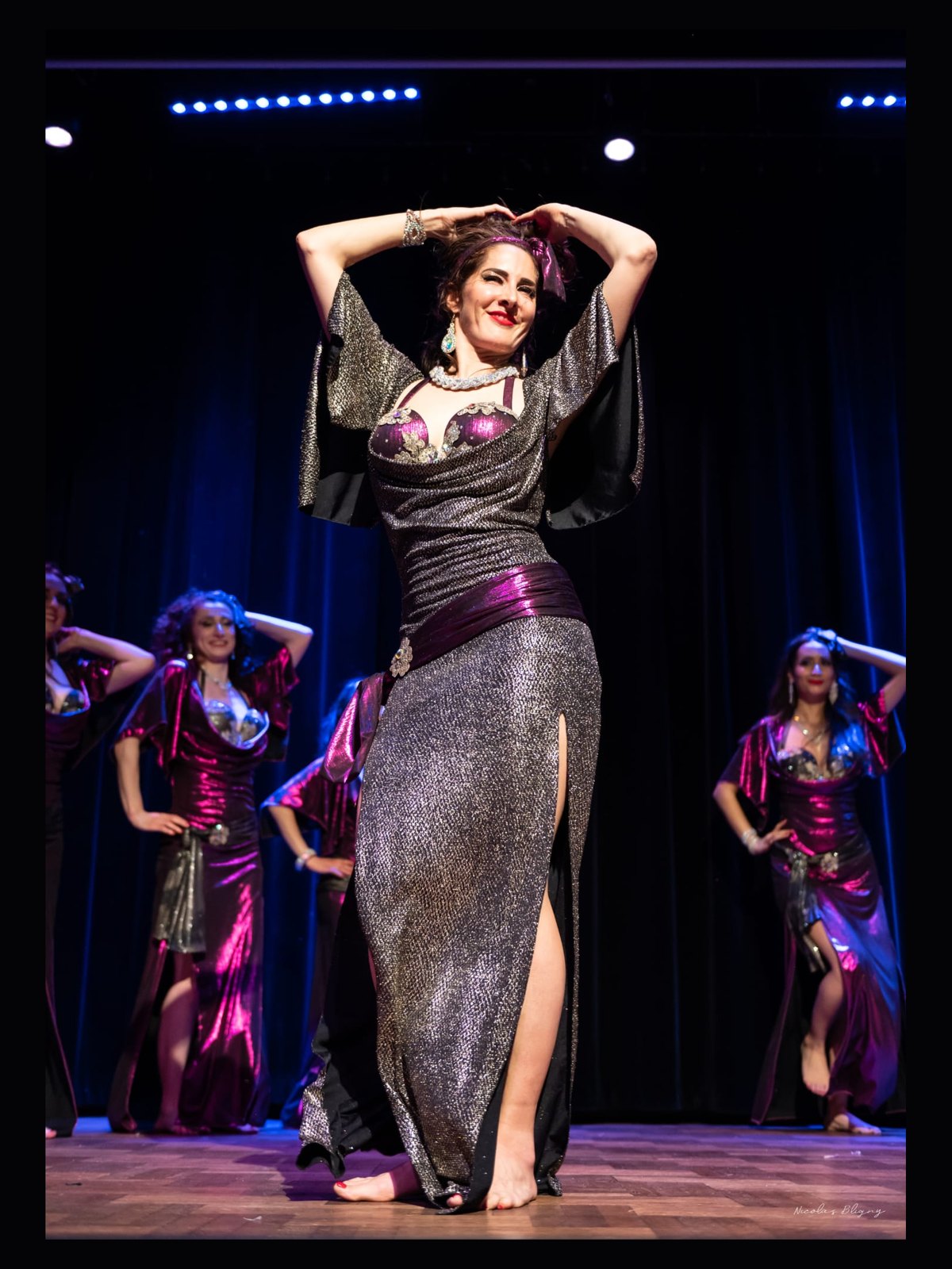 Ciya en robe argentée et violette, mains sur la tête, devant les danseuses de sa compagnie